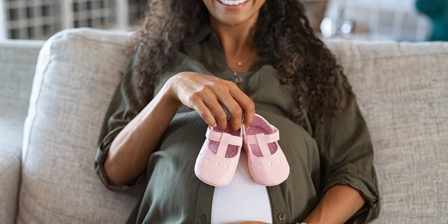 Pregnant woman sits on the couch and holds a pair of baby shoes in front of her baby bump.