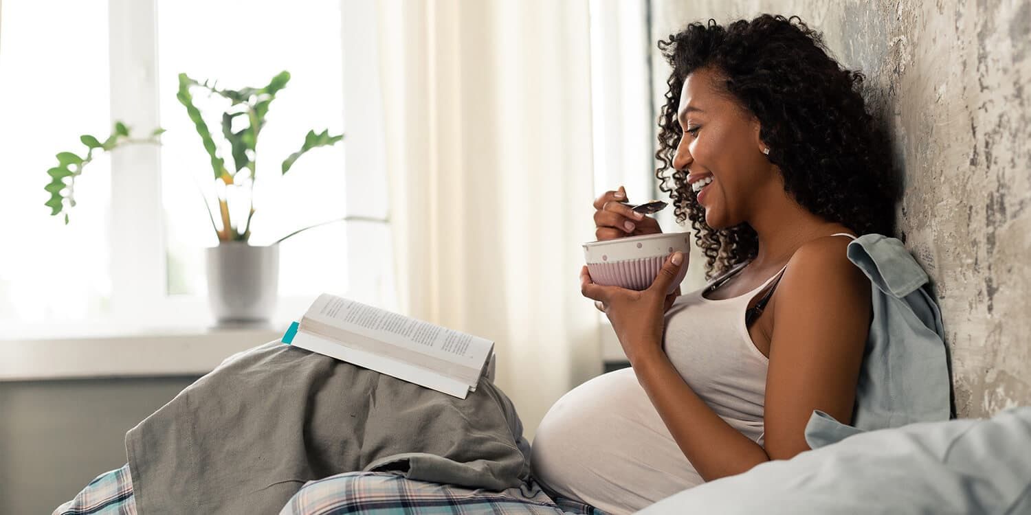 Een lachende, zwangere vrouw zit met ontbijt op bed.