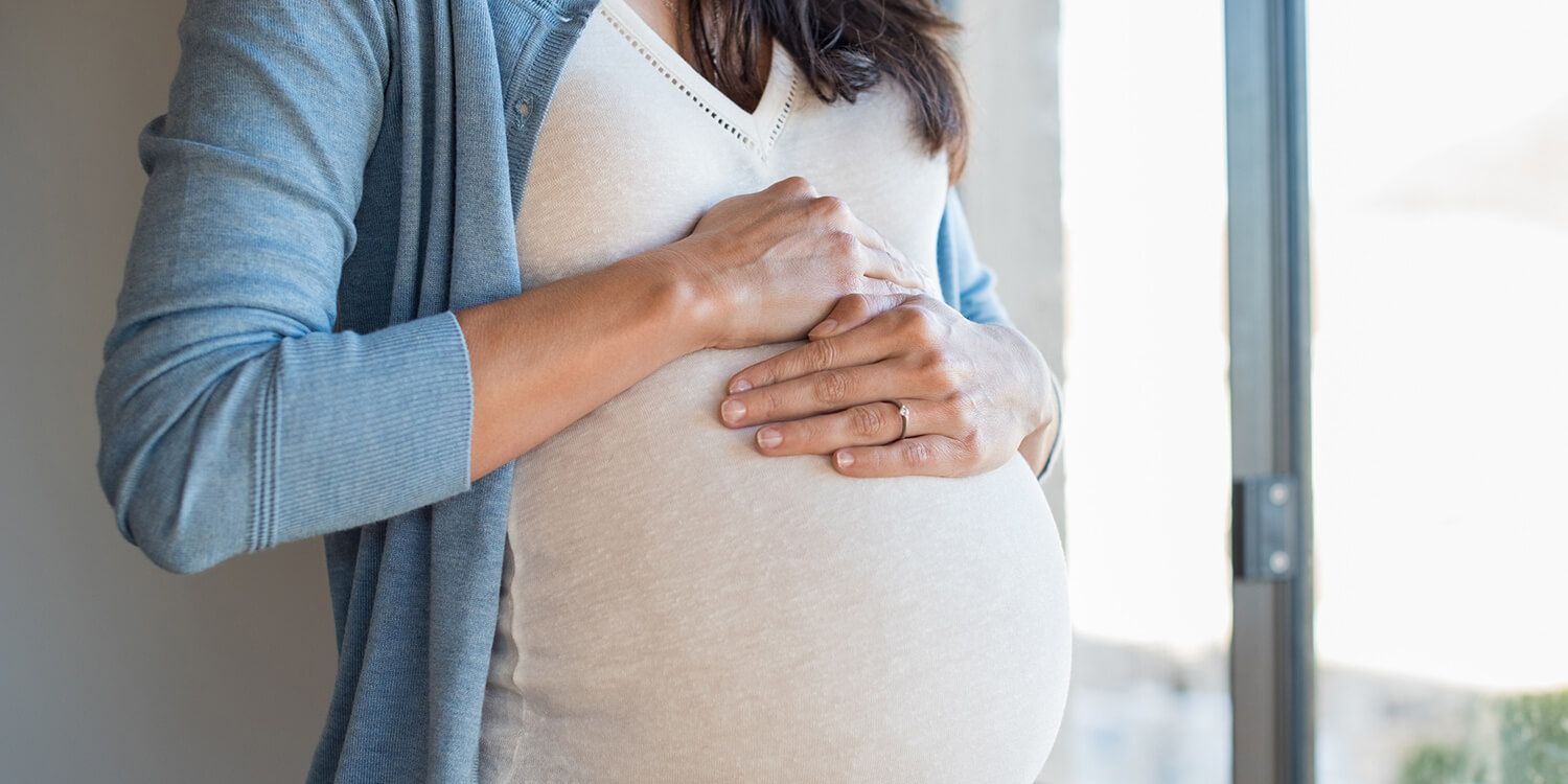 Gravid kvinde lægger hænderne på maven og ser ud af vinduet