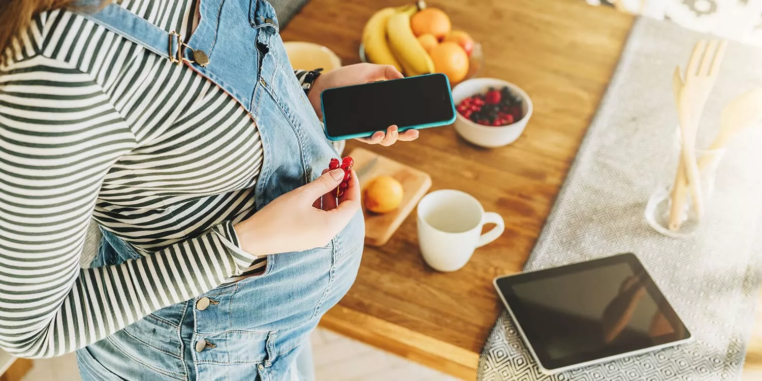 mulher grávida segurando o celular em uma das mãos e frutas na outra, ao fundo há uma mesa de jantar com frutas.