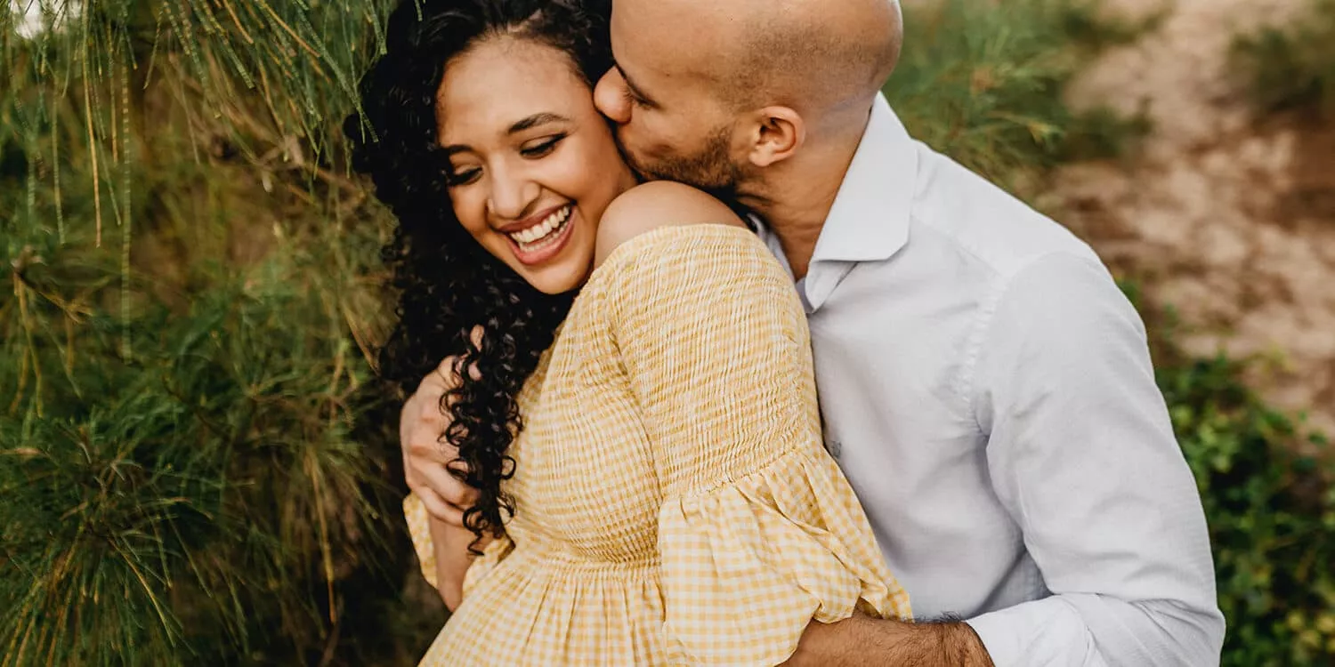 Man has his arms around his pregnant partner and is kissing her on the cheek 