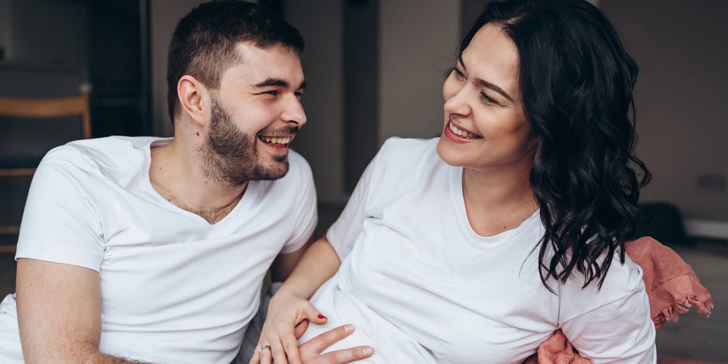 Junges schwangeres Paar kuschelt auf Couch, beide legen ihre Hände auf Babybauch der Frau. (14. SSW)