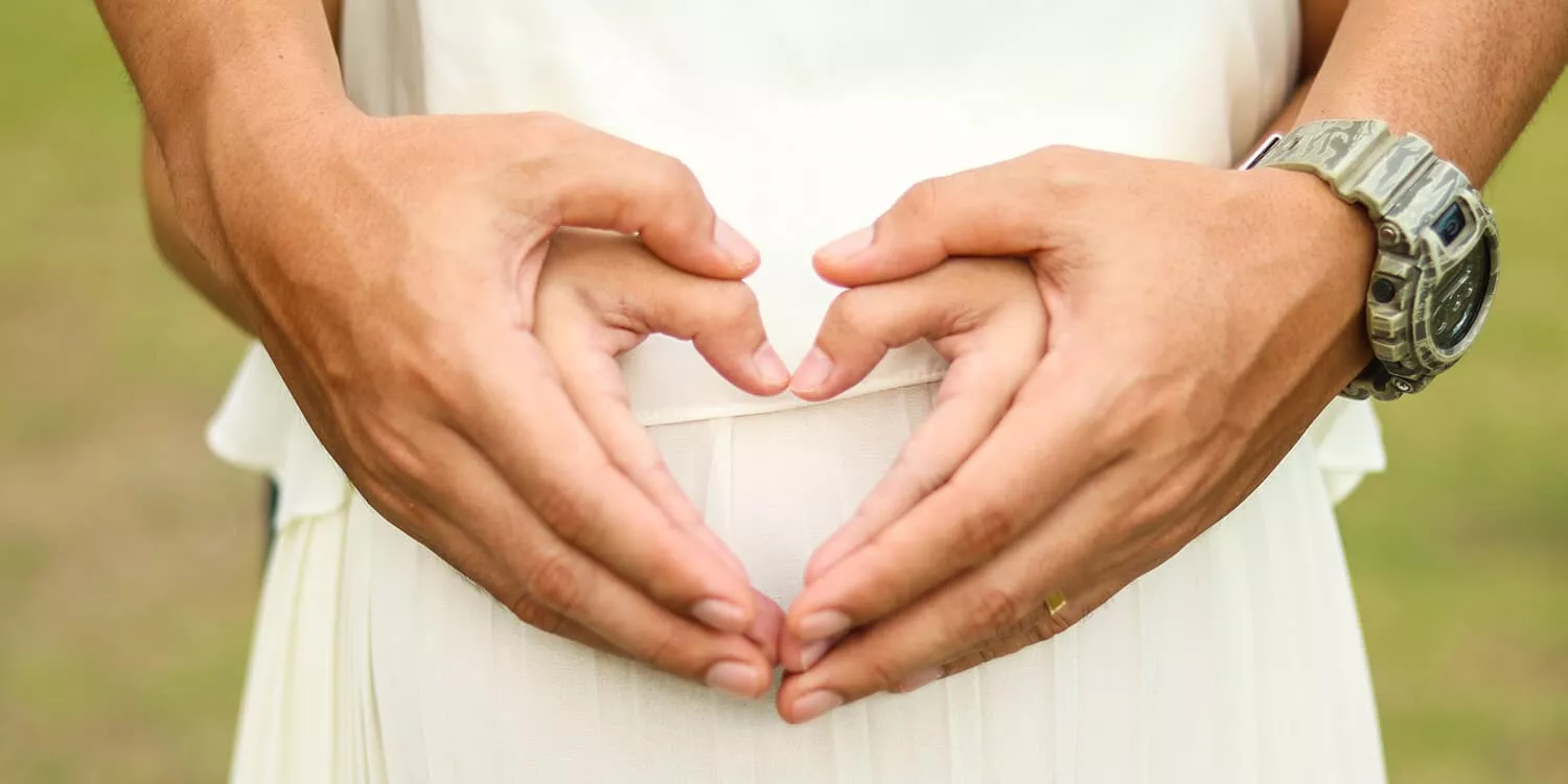 A couple forms the shape of a heart with their hands in front of the woman's stomach