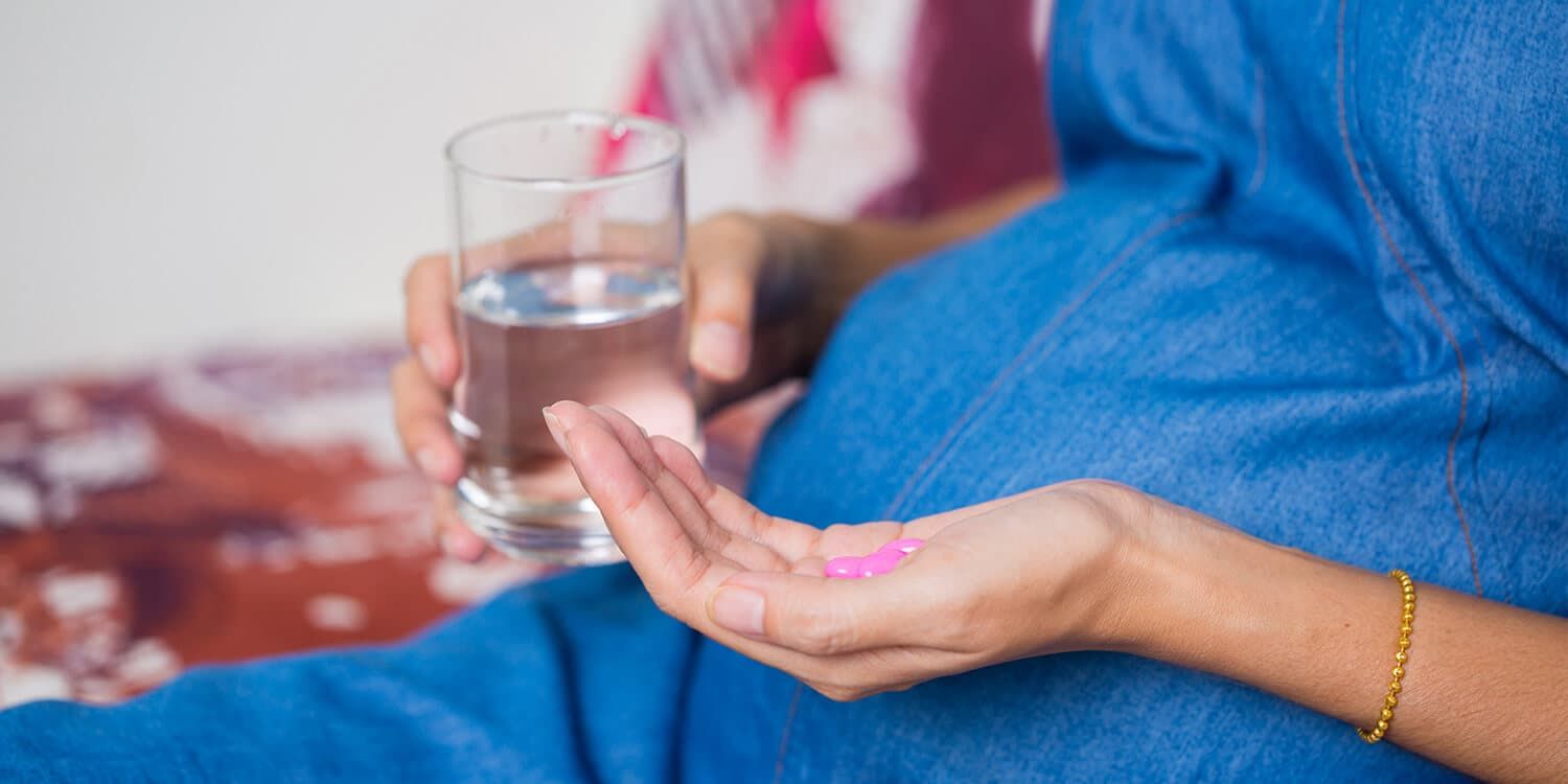 Pregnant woman sits on a couch and holds pills and a glass of water in her hands. 