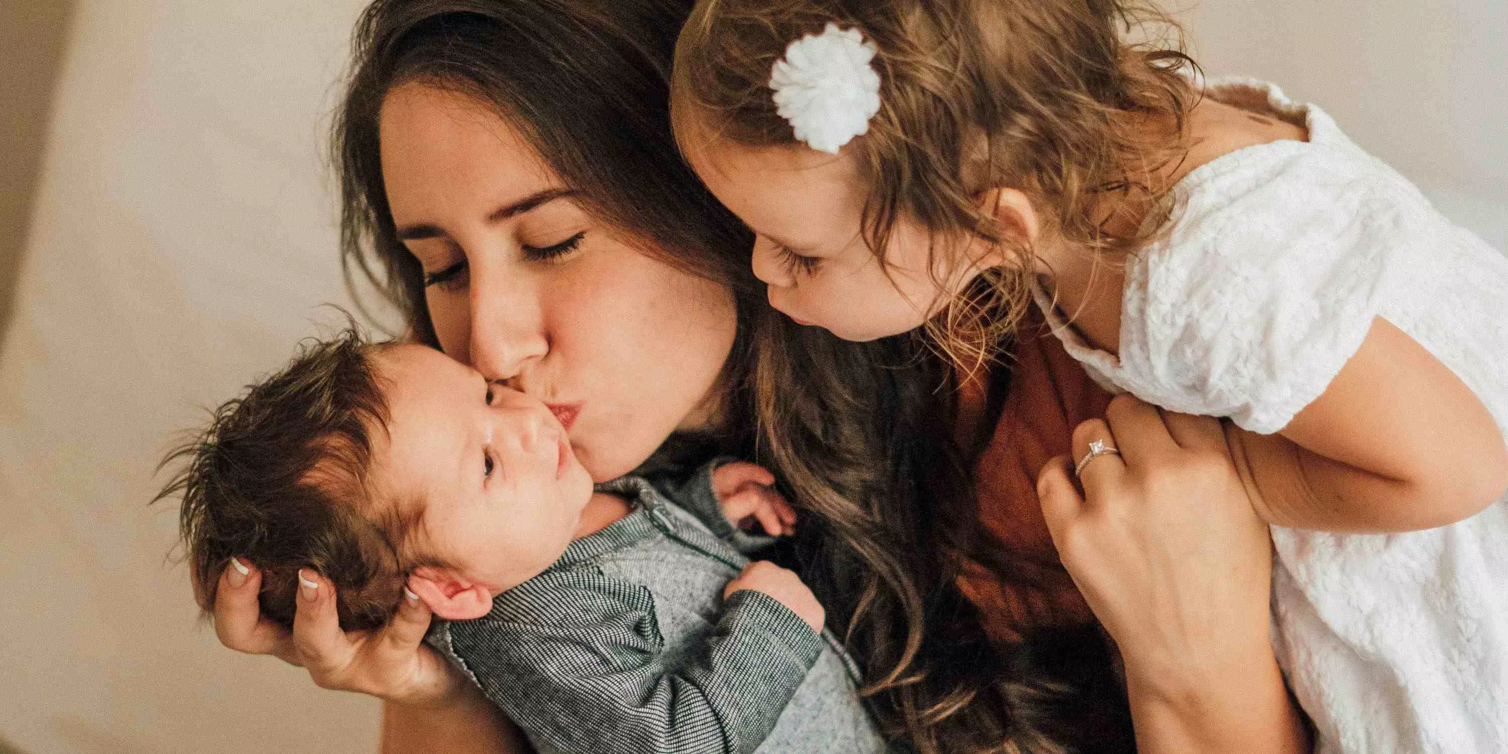Mutter hält das Neugeborene im Arm und küsst es auf die Wange, das Geschwisterkind steht neben der Mutter und haucht dem Baby einen Kuss zu