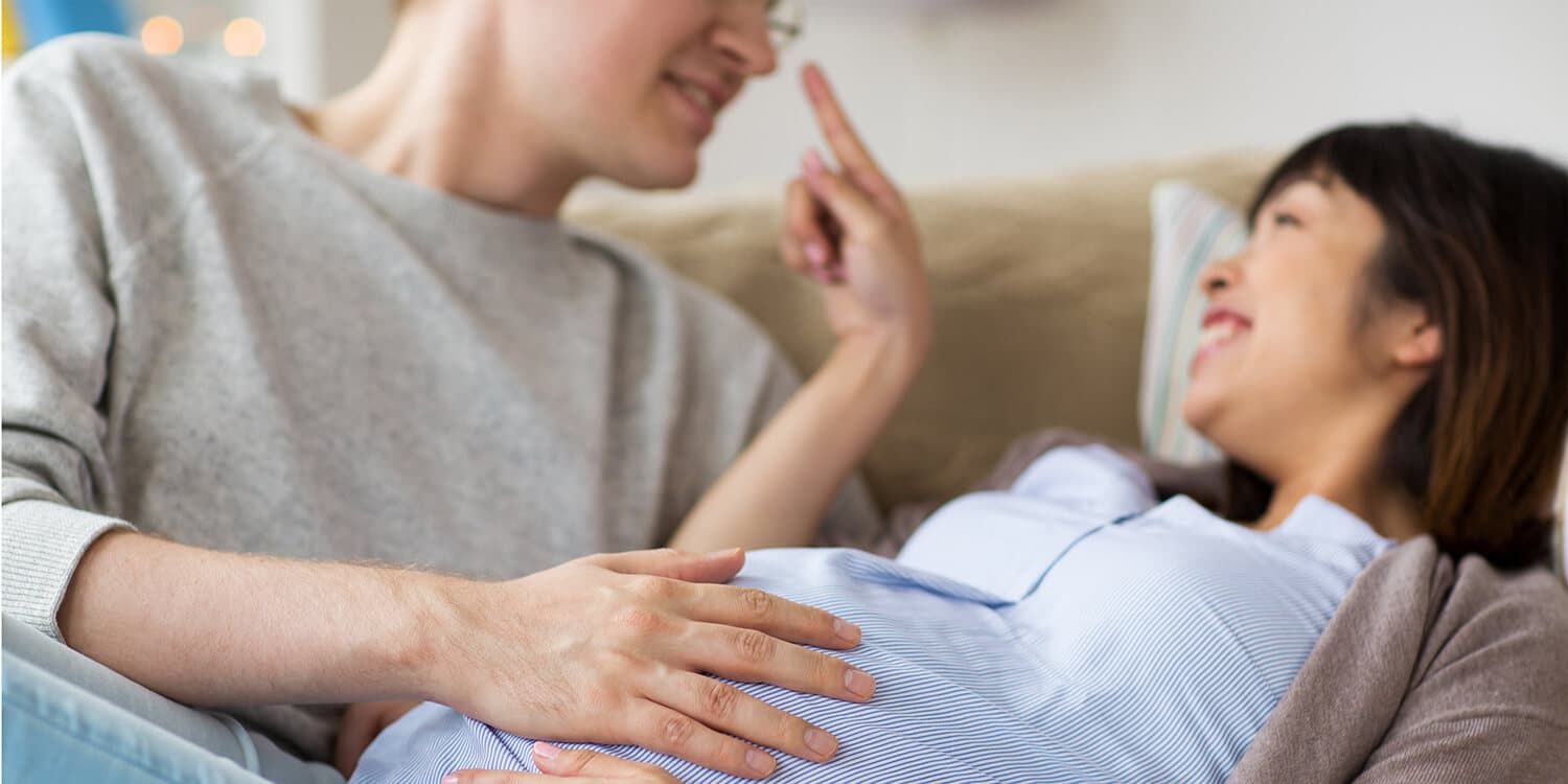 Una pareja joven está acurrucada en el sofá y riéndose; el hombre pone las manos en la barriguita de la mujer.