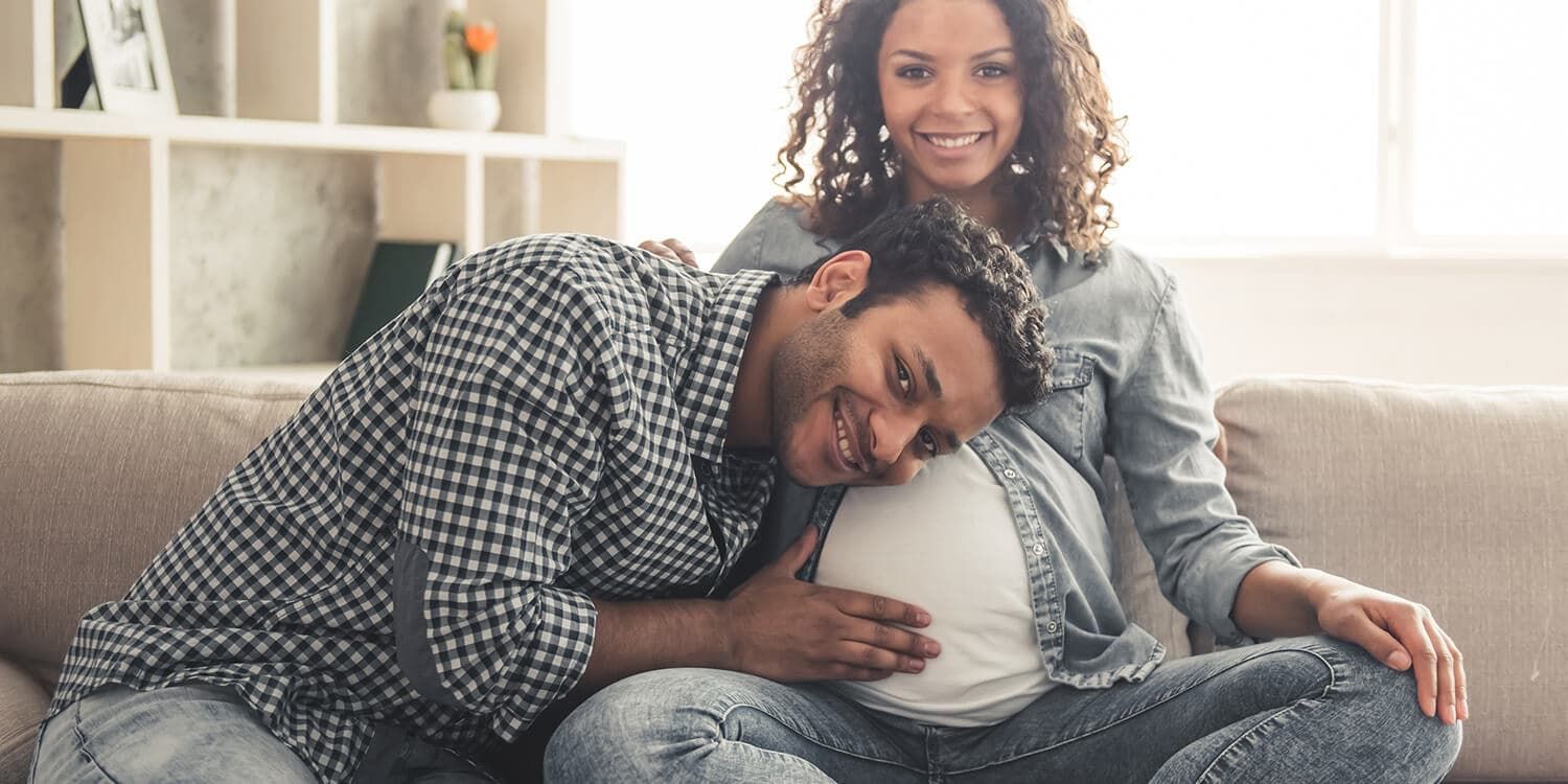 Mann und schwangere Frau sitzen auf Sofa, Mann hält Ohr an Bauch von Frau