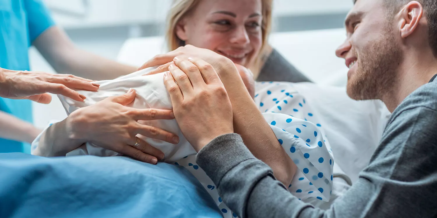 In hospital, a midwife is giving the newborn baby to the mother to hold, a supportive father is lovingly stroking the baby's head. 