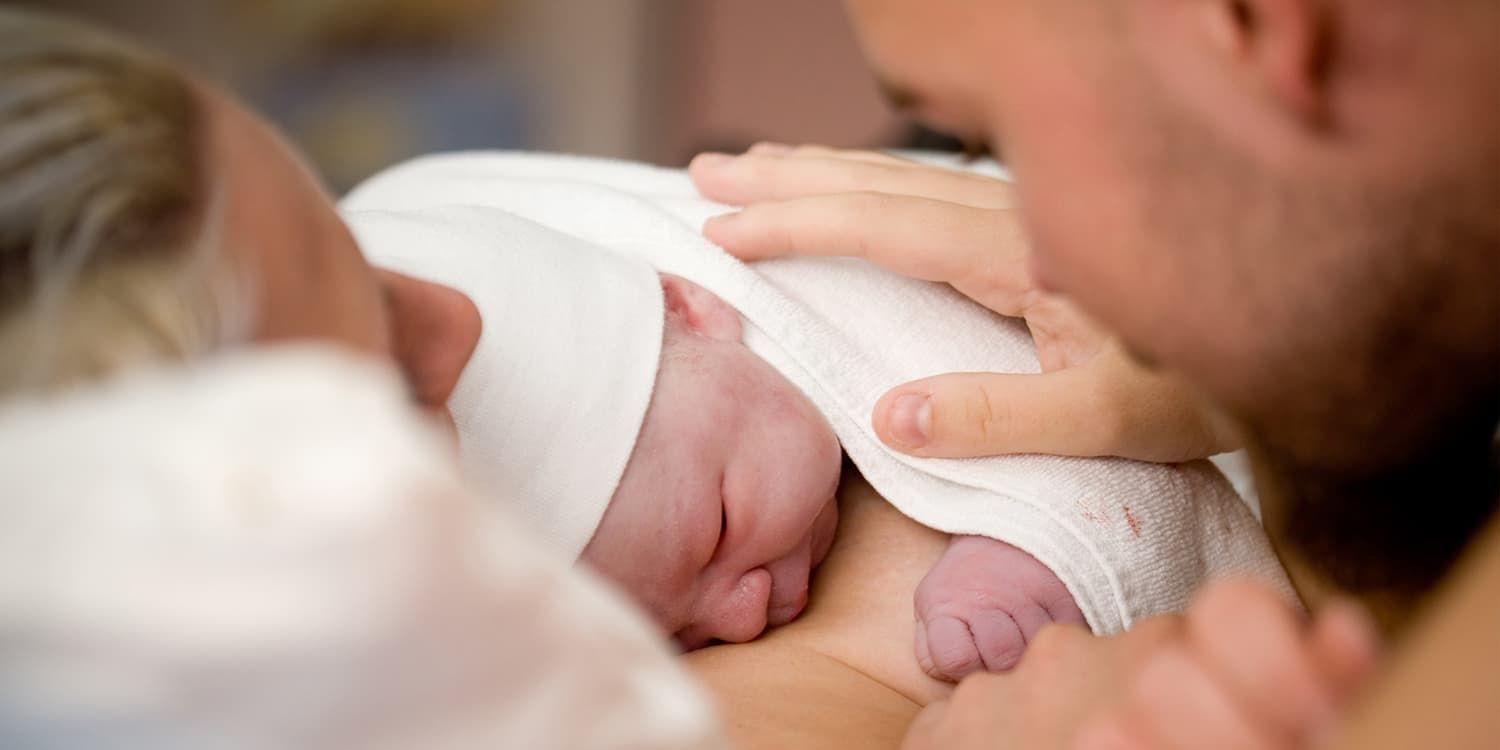 Een pasgeboren baby ligt op moeders borst en de vader legt liefdevol zijn hand op de baby.