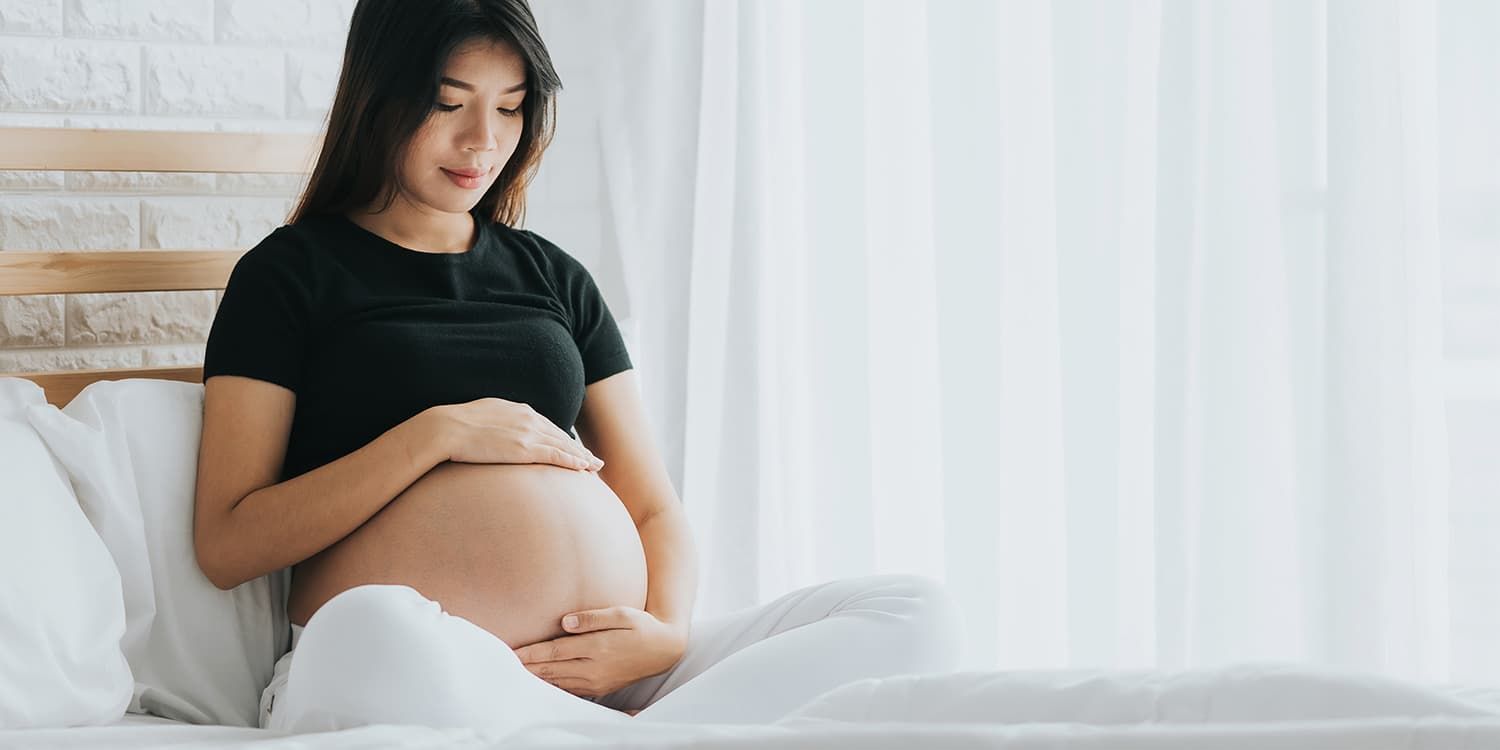 Schwangere Frau sitzt auf Bett und umgreift vorsichtig ihren Babybauch.