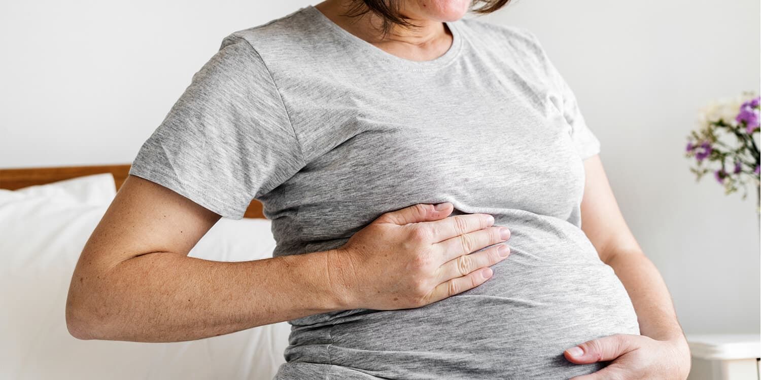 Uma mulher grávida sentada na cama coloca as mãos sobre a sua barriga e sorri