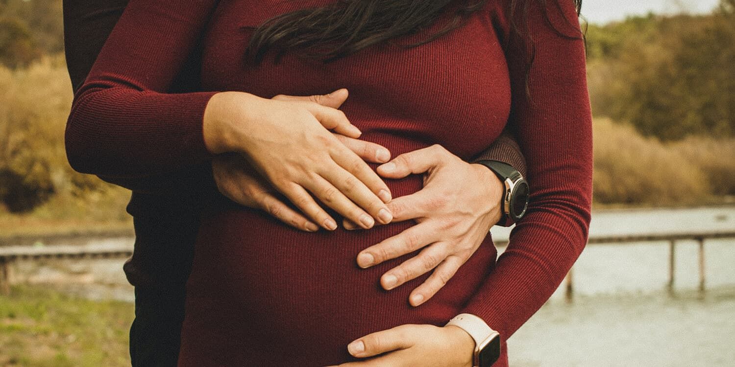 Coppia in attesa si sofferma all'aperto, entrambi abbracciano il pancione della donna 