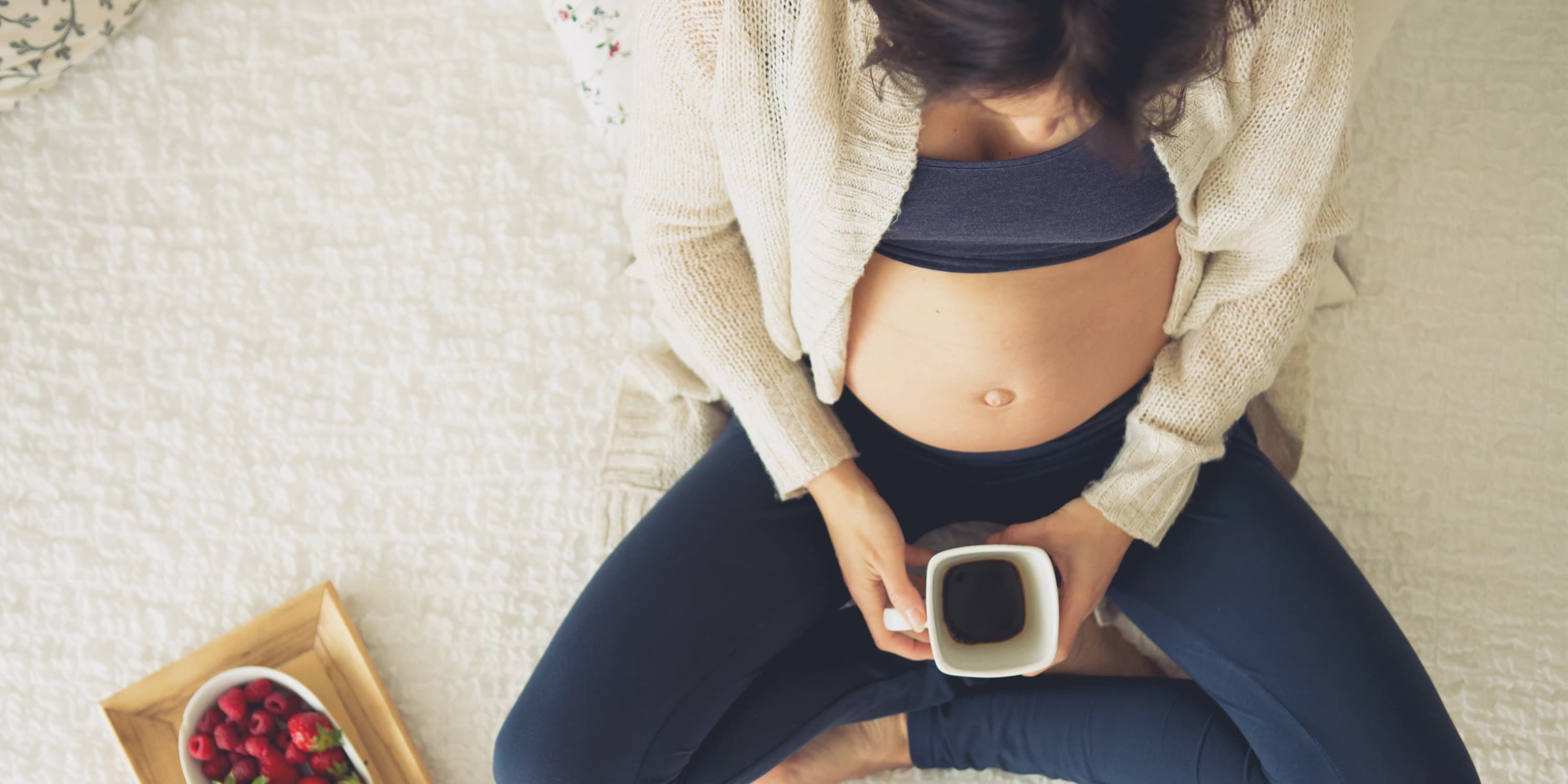 Uma mulher grávida está sentada no chão com uma chávena de café e morangos num tabuleiro