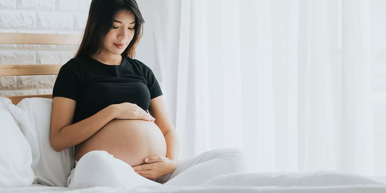 Gravid kvinde sidder på sengen og omfavner ømt sin mave.