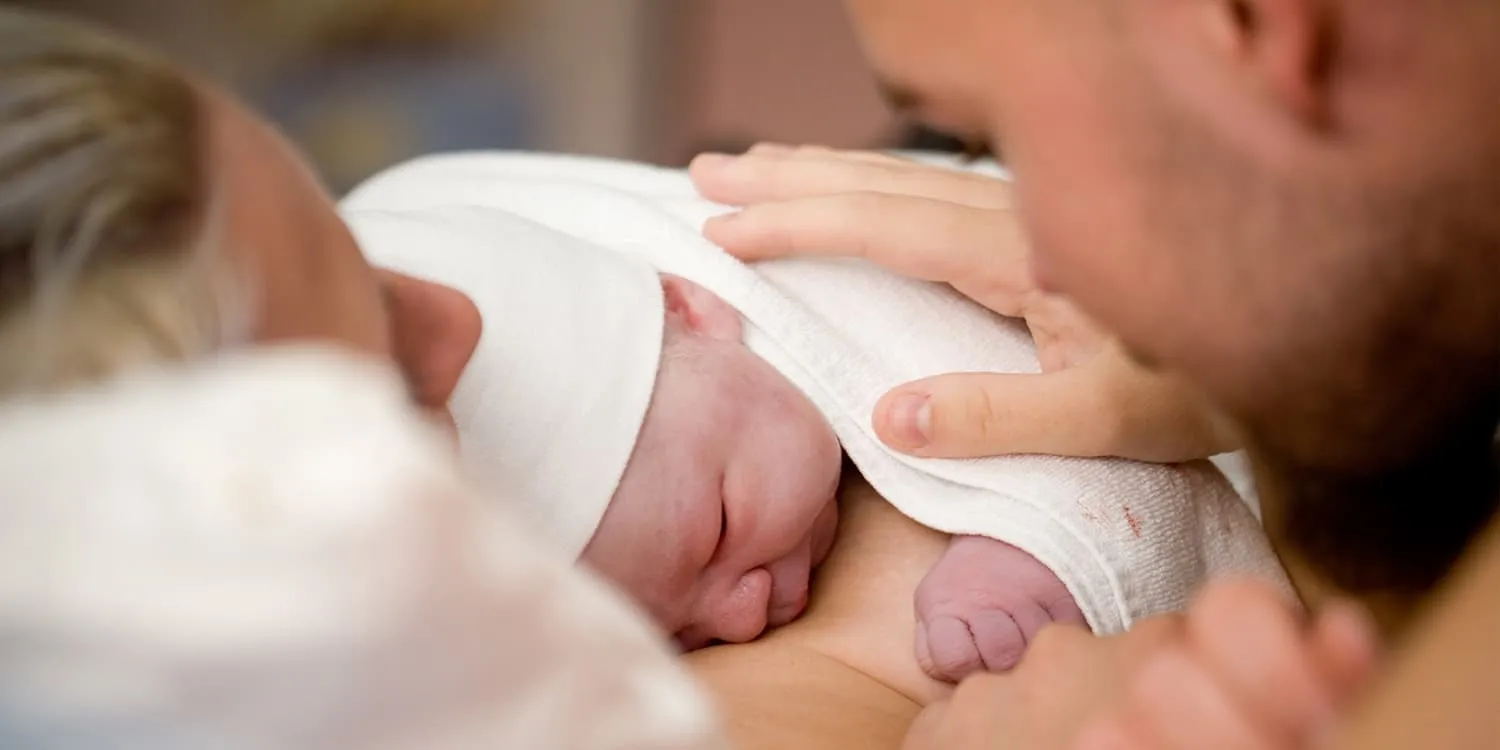 Nyfødt baby ligger på mors bryst, far legger hånden kjærlig på babyen