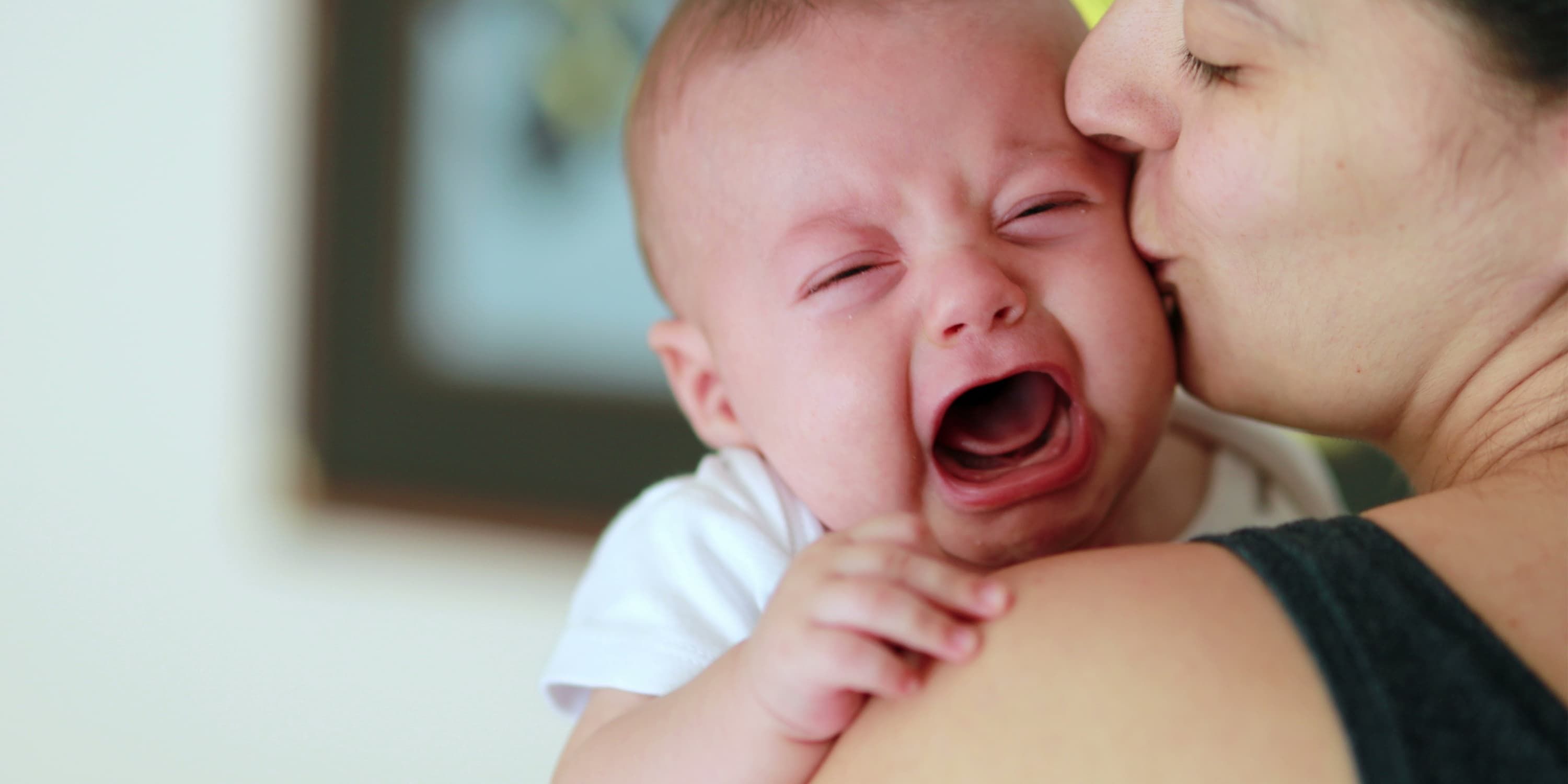 Mother carries her crying baby over her shoulder and kisses it on the cheek.