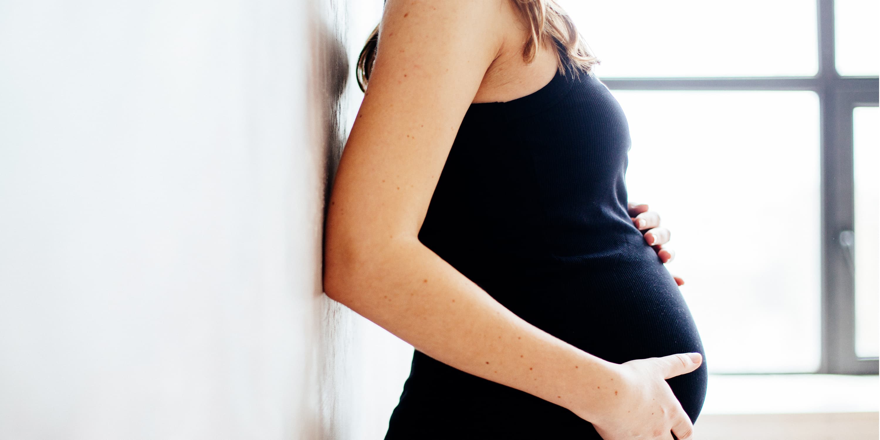 Side view, pregnant woman leans against the wall and puts hands on her belly
