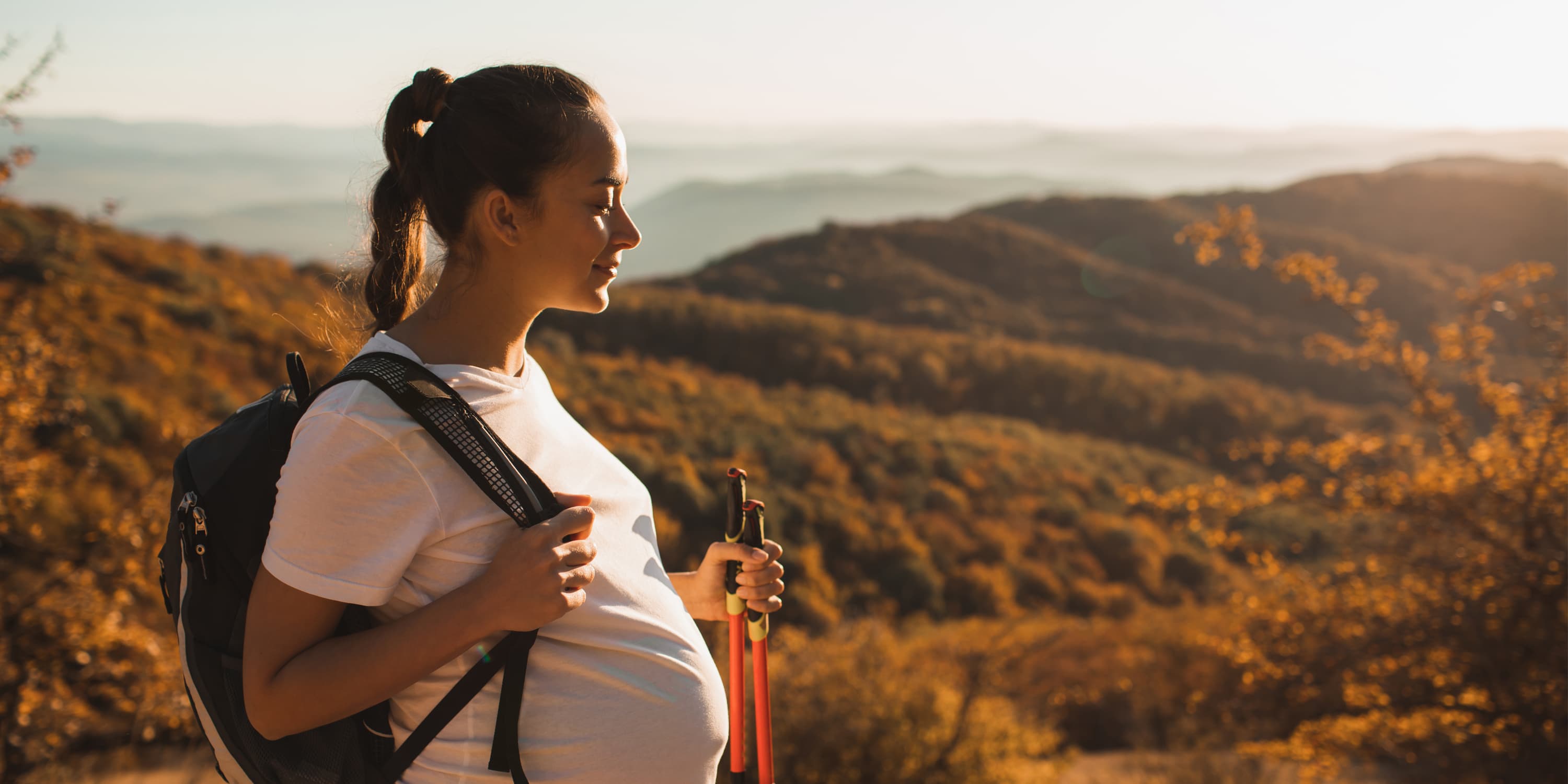 Seitenansicht einer schwangeren Frau beim Wandern mit Wanderstöcken und Rucksack.