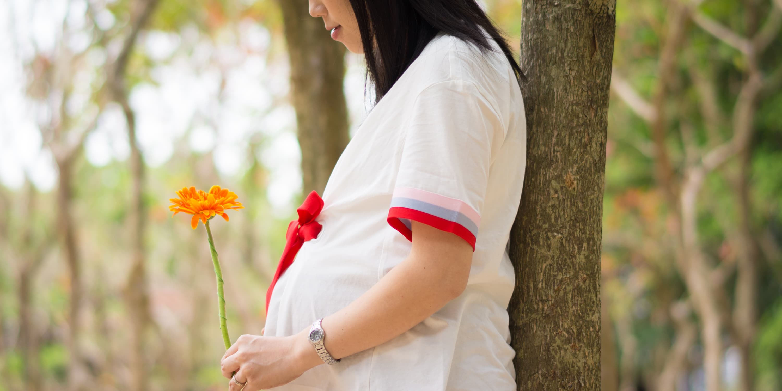 Seitenansicht: Schwangere Frau steht unter einem Baum und hält eine Blume in den Händen