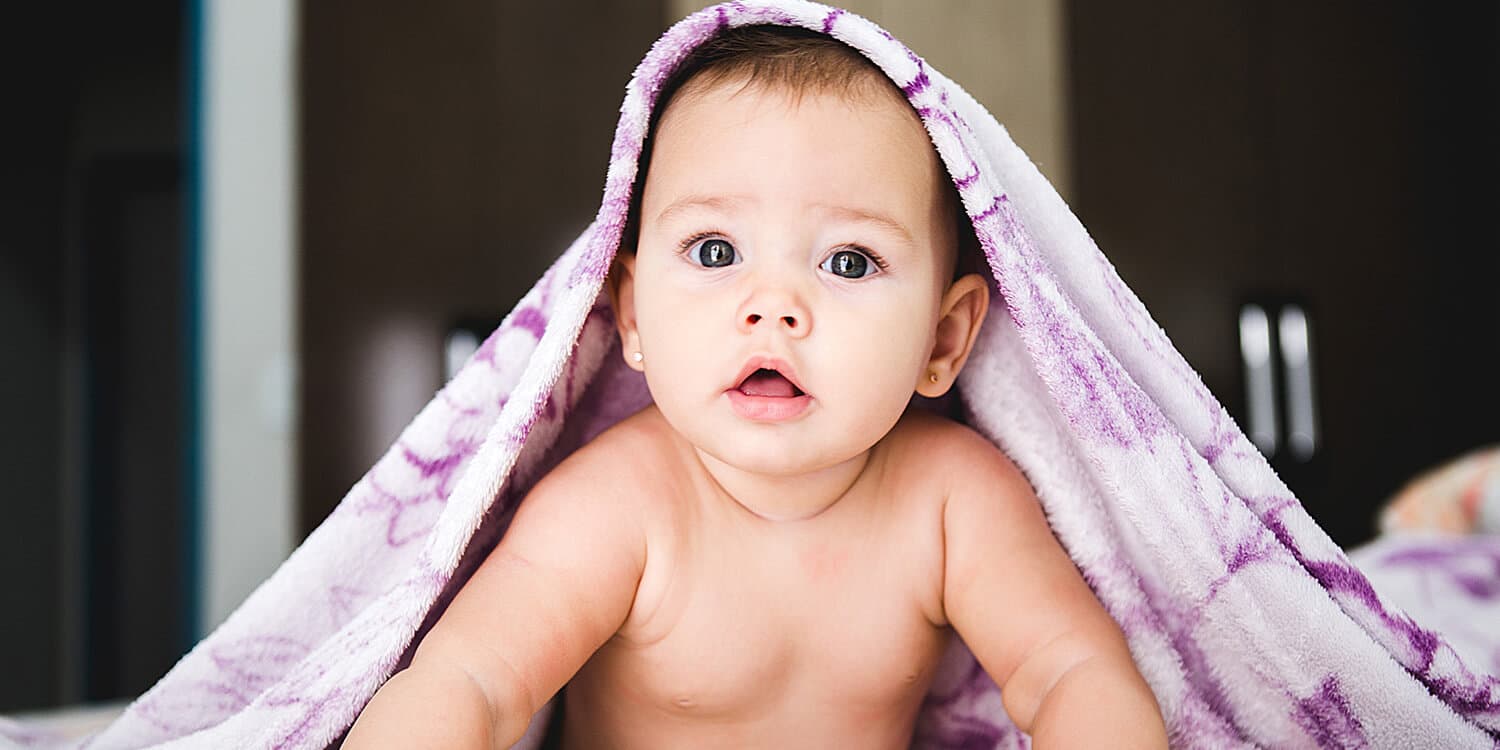 Baby lying on stomach with towel over head and looking at camera