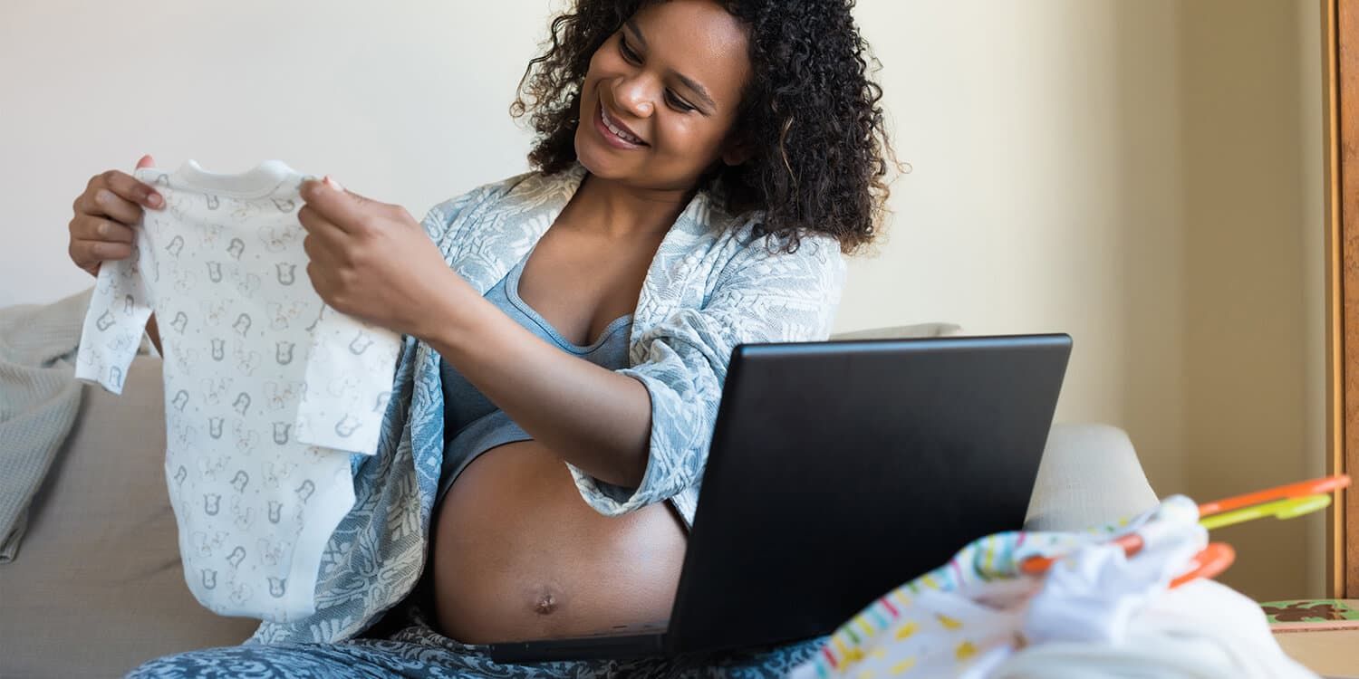 Frau sitzt auf Sofa und shoppt online für ihr Baby. Dabei betrachtet sie einen Baby-Strampler, den sie in den Händen hält (27. SSW)