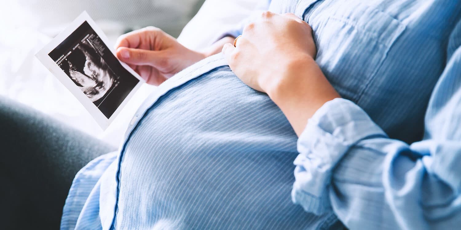 Schwangere Frau hält Ultraschallbild in der Hand und greift sich mit der anderen Hand auf den Bauch (20. SSW)